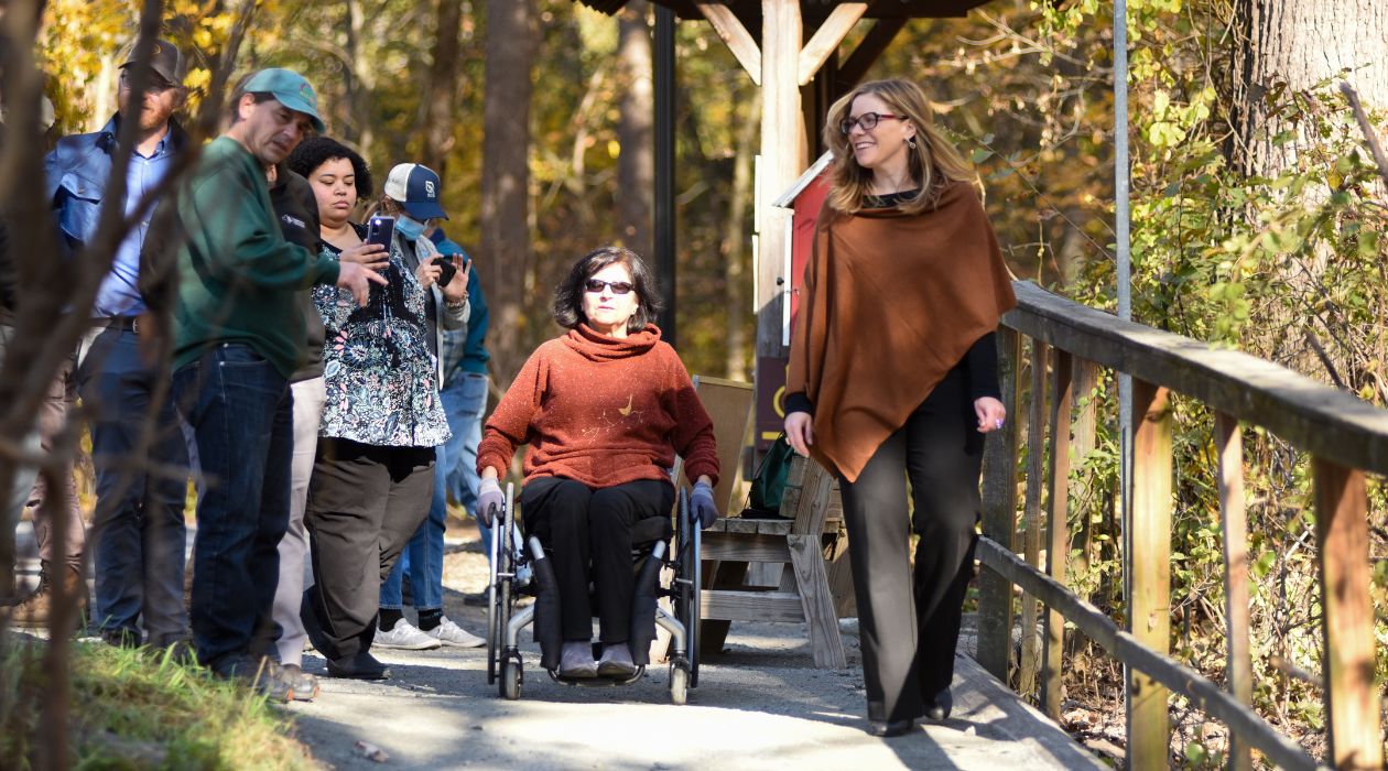 A woman in a wheelchair with shoulder length dark hair, sunglasses, a rust colored sweater and black pants makes her way along the Woodland Trail at Stony Kill Farm in Wappingers Falls accompanied by several other adults.