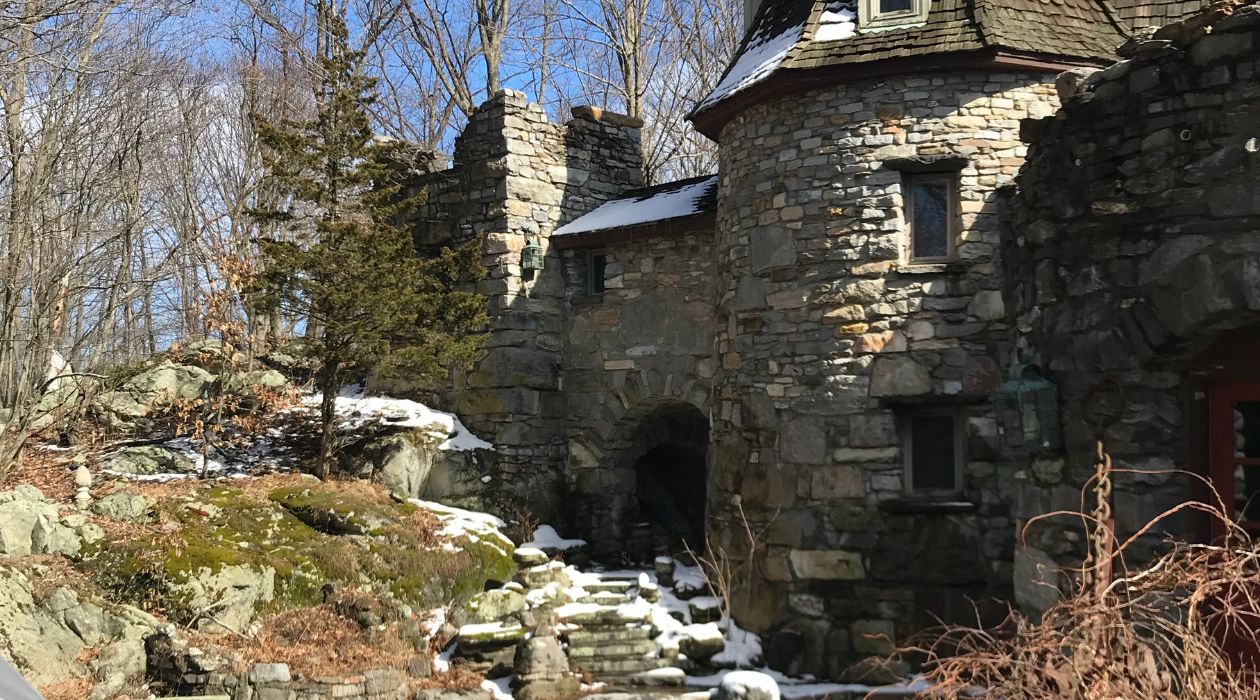Wings Castle in Millbrook is a stone castle with a wood shingle roof atop its tower. It was photographed on a winter day with a light dusting of snow with moss covered rocks in the foreground and trees with no leaves in the background.