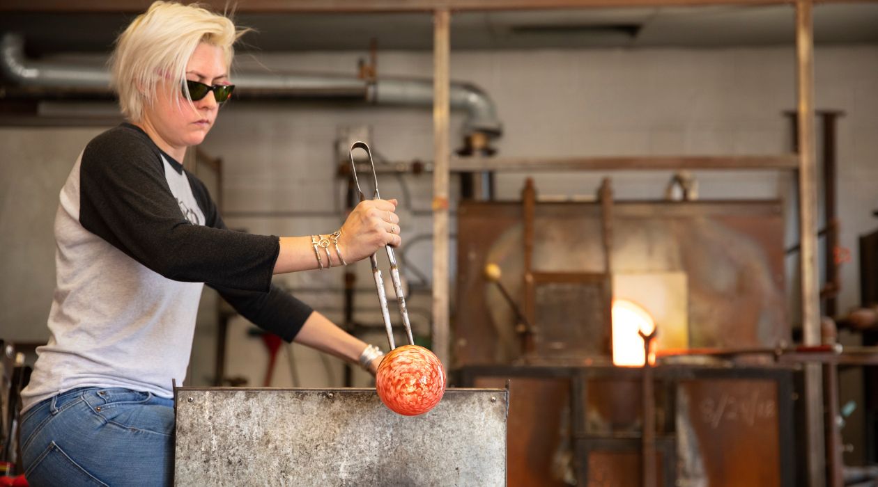 A female with blonde hair, sunglasses, blue jeans and a black and gray tee shirt uses metal tools to prepare an orange glass ornament at Hudson Beach Glass, Beacon